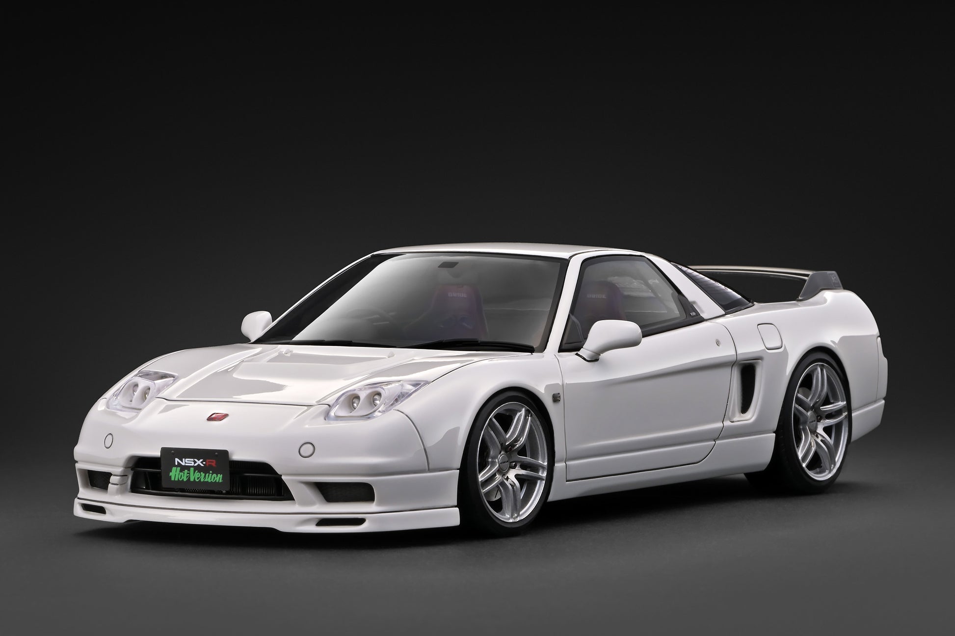 A white widebody sports car with a black soft top, shown at a three-quarter front angle on a dark gray studio background. The car features custom silver multi-spoke wheels, a front lip spoiler, and a green and white license plate frame.