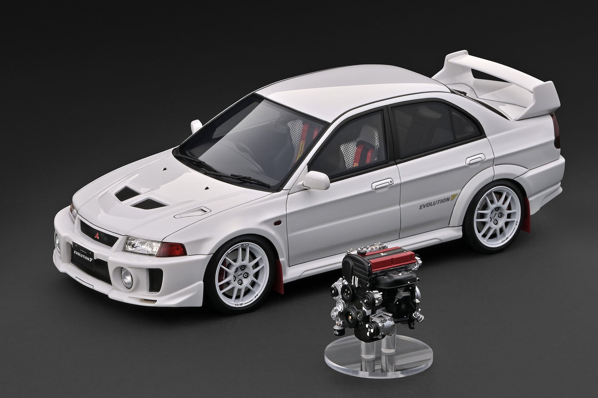 A white model car with a large rear spoiler and a small engine display stand in front, photographed on a dark gray studio background.