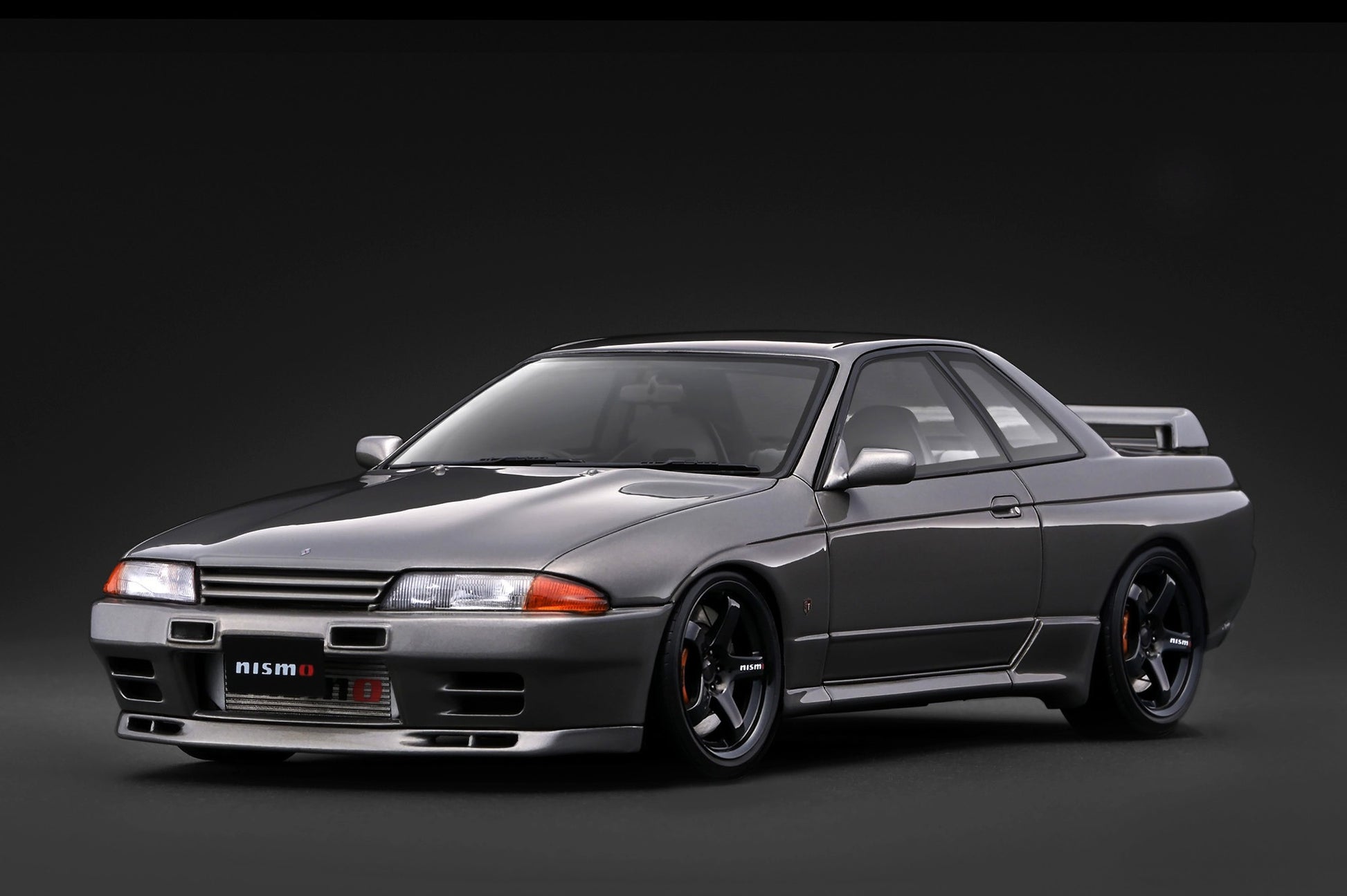 A dark gray Nissan R32 GT coupe with a black hood and custom black wheels, photographed from a three-quarter front angle against a dark studio background.