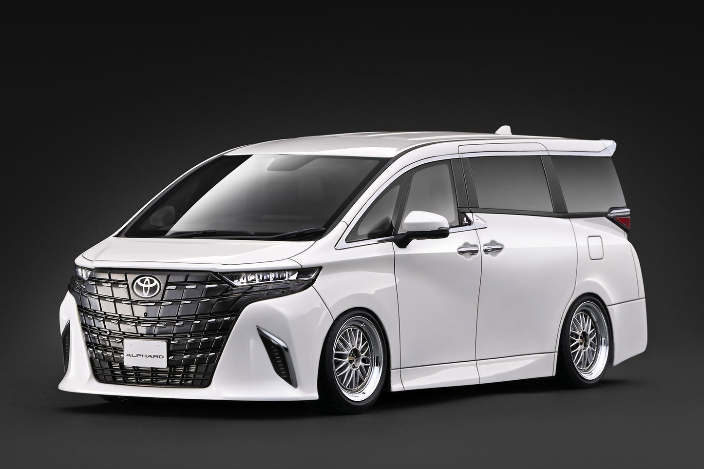 A white customized Toyota Alphard minivan with a black front grille and multi-spoke alloy wheels, photographed on a dark studio background.