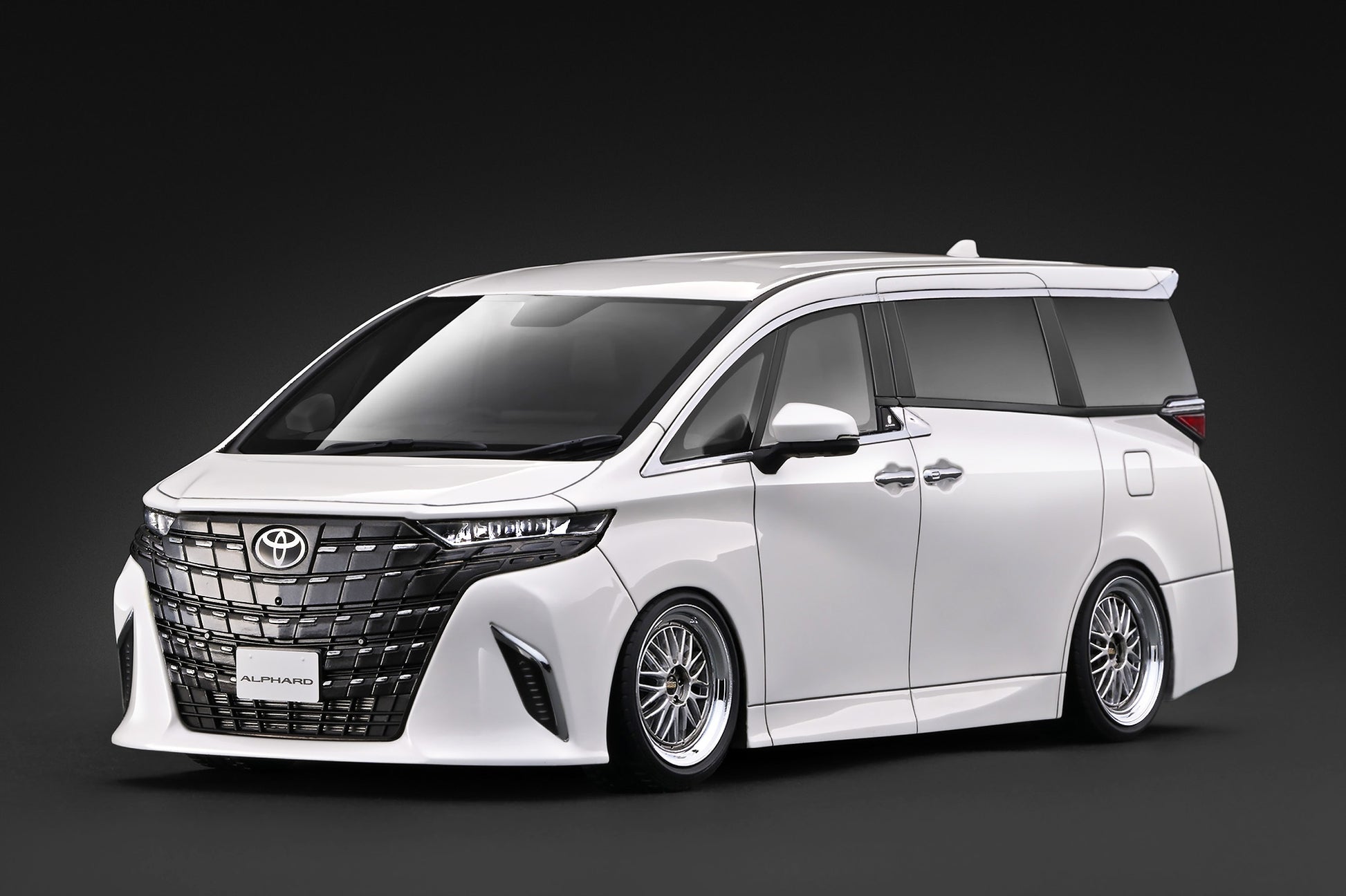 A white customized Toyota Alphard minivan with a black front grille and multi-spoke alloy wheels, photographed on a dark studio background.