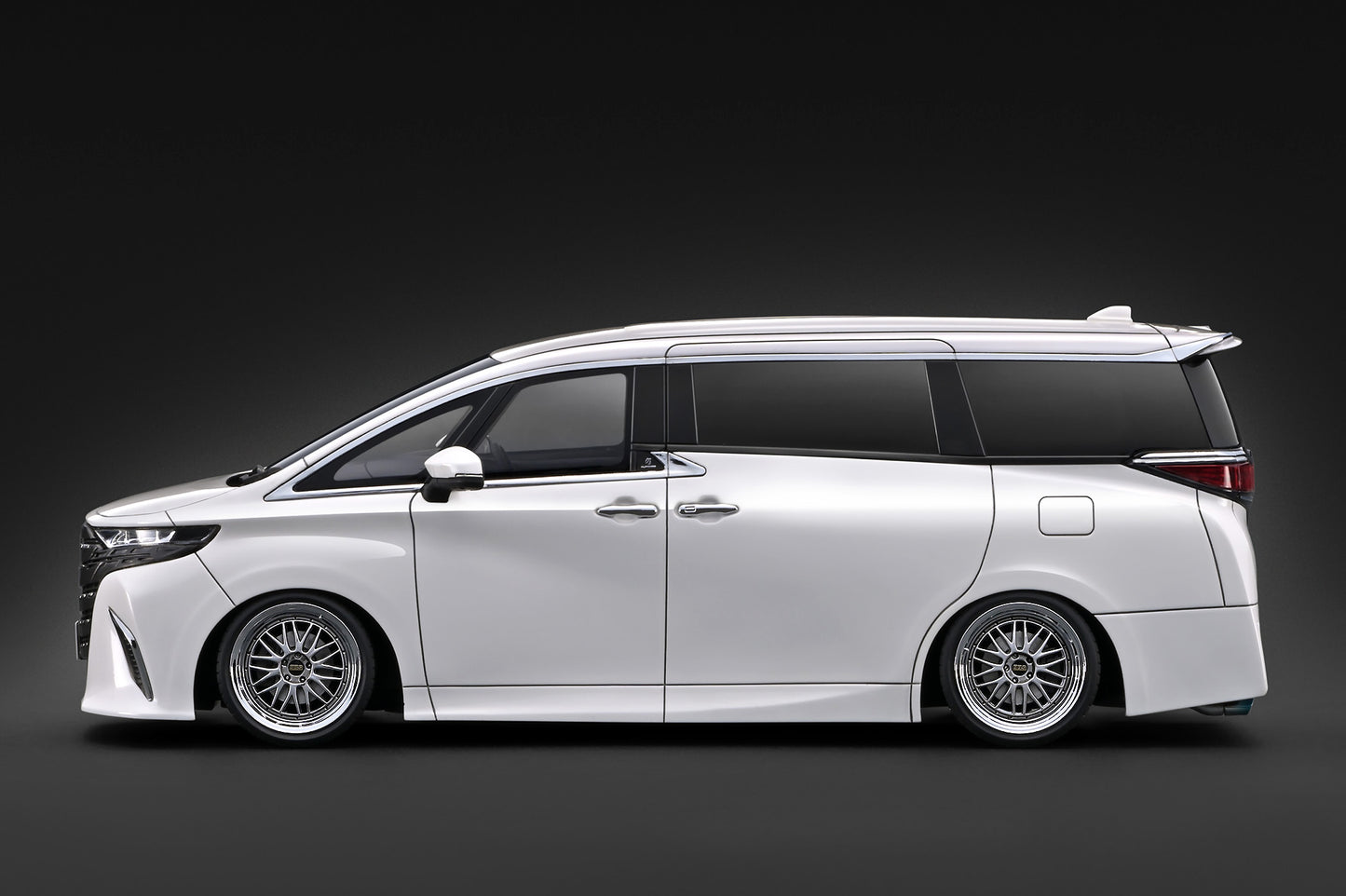 A white customized Toyota Alphard van with a lowered suspension, custom multi-spoke wheels, and a rear spoiler, shown in a side profile against a dark studio background.