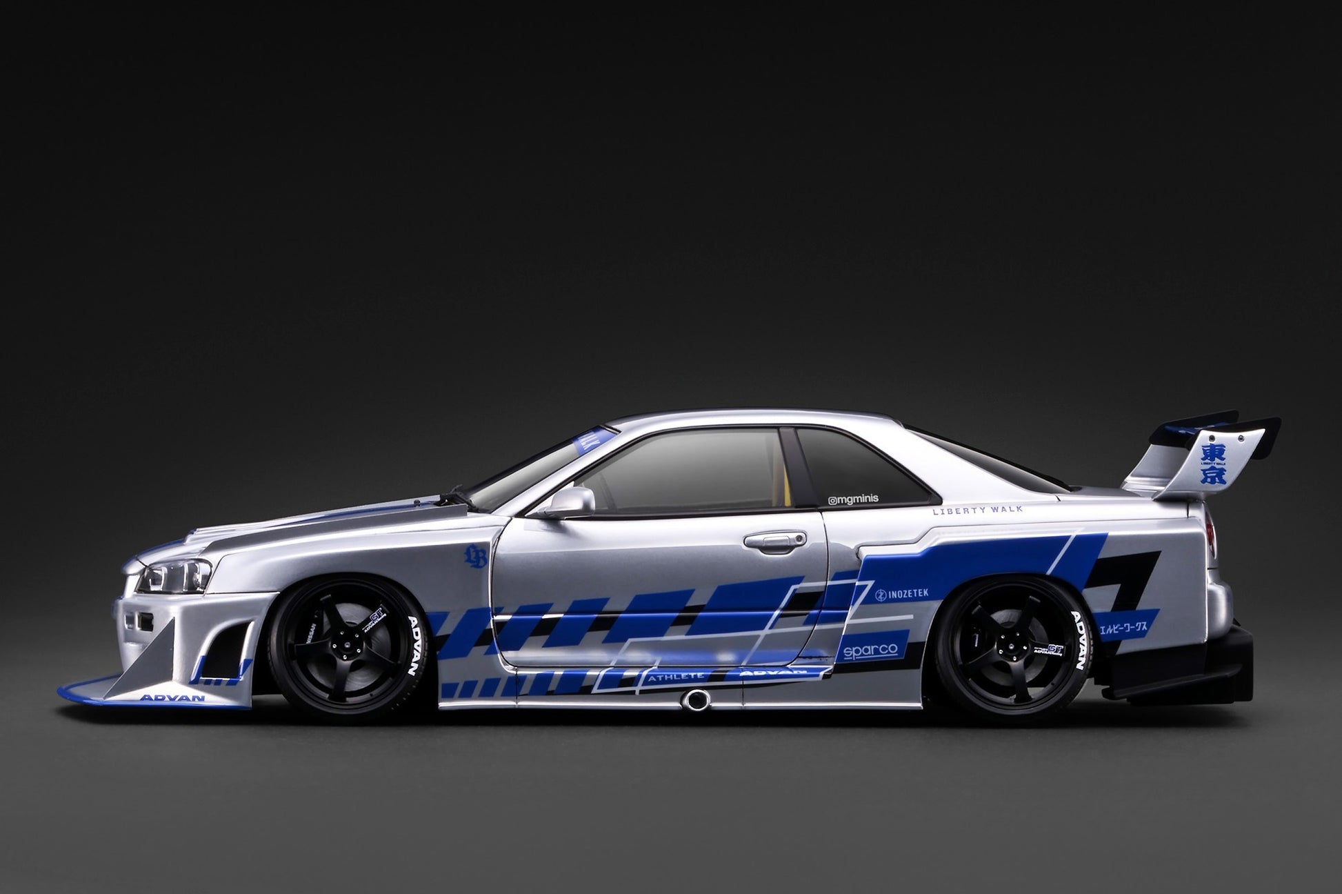A silver Nissan R32 GT race car with blue and black graphics, shown in a side profile against a dark studio background.