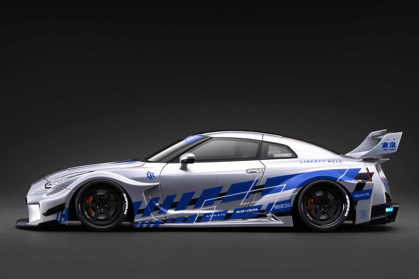 A silver and blue widebody sports car with a large rear wing and racing decals, shown in a clean side profile against a dark studio background.