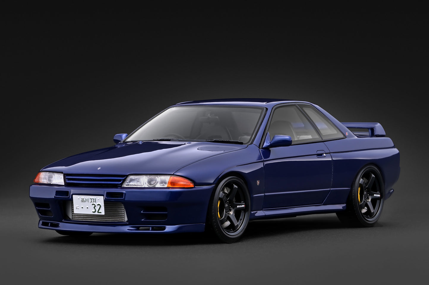 A dark blue Nissan Skyline R32 sports coupe with black wheels and yellow brake calipers, photographed on a dark gray studio background.