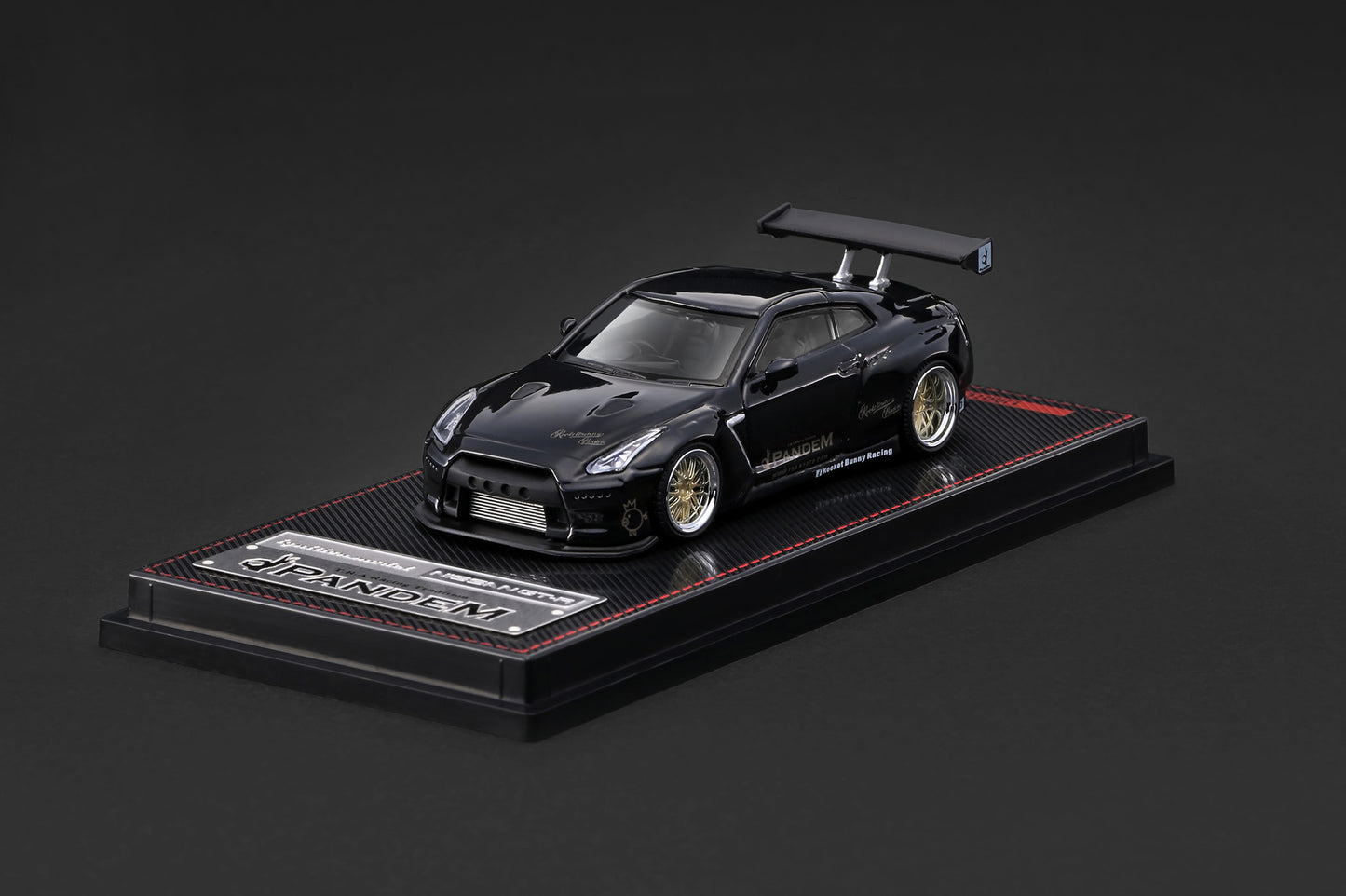 A black Nissan GT-R diecast model car displayed on a black base with a carbon-fiber texture and red stitching, set against a dark gray background.