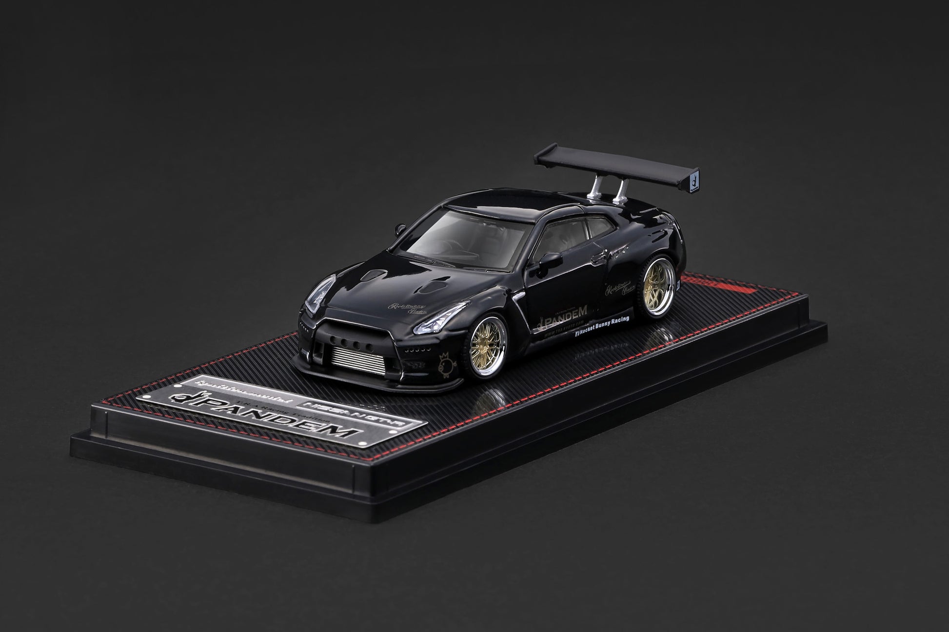 A black Nissan GT-R diecast model car displayed on a black base with a carbon-fiber texture and red stitching, set against a dark gray background.