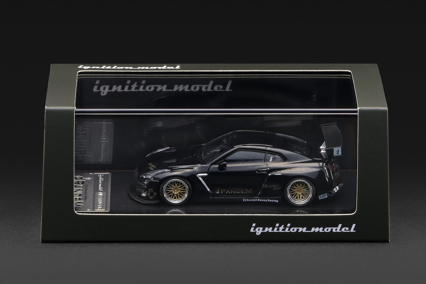 A black die-cast model car in its original display box, featuring a Nissan R35 GT-R with gold wheels and sponsor decals. The packaging is branded with 'ignition model'.