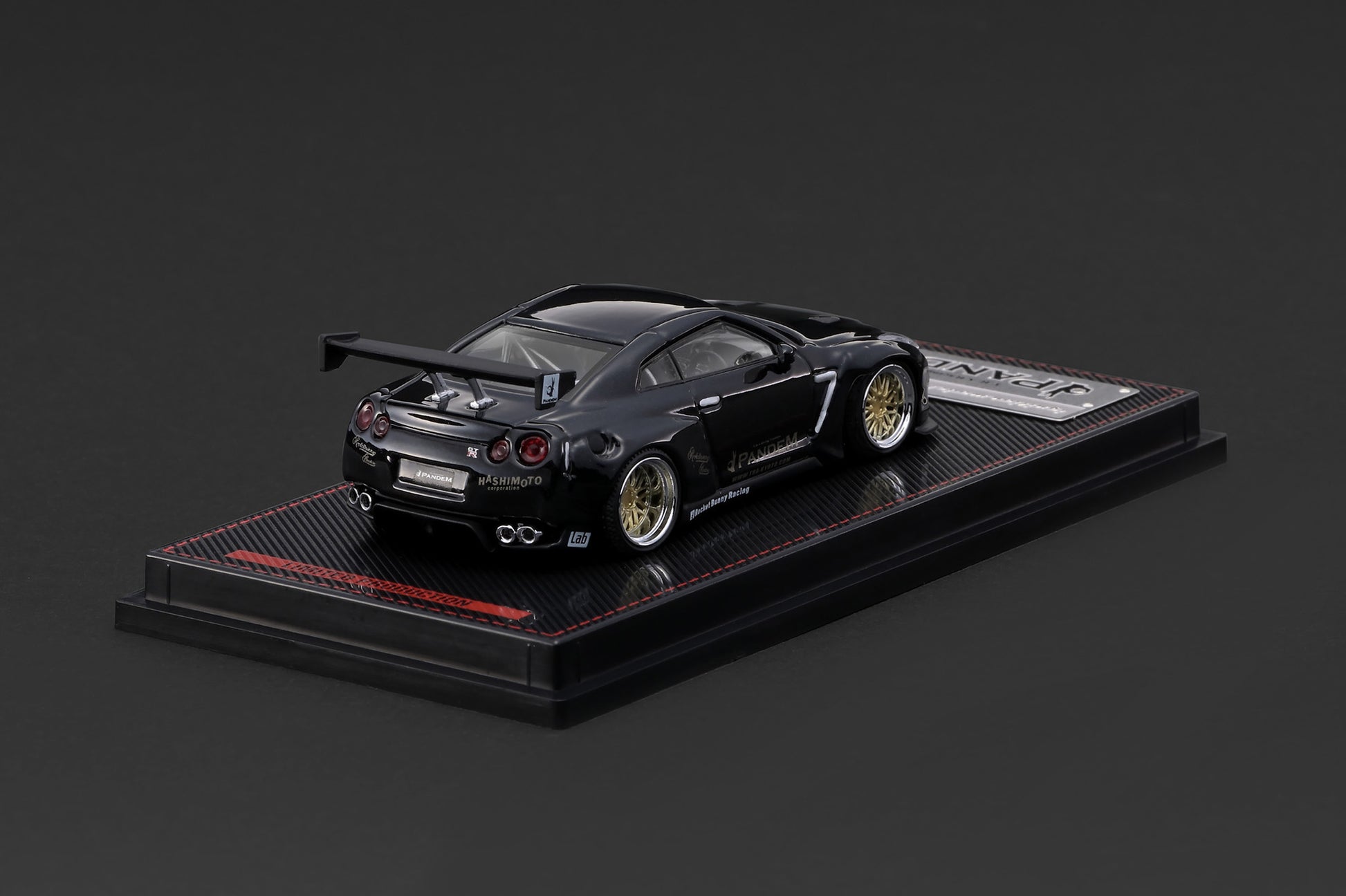 A black die-cast model of a Nissan GT-R Nismo race car, displayed on a black textured base with a small silver plaque, set against a dark gray background.