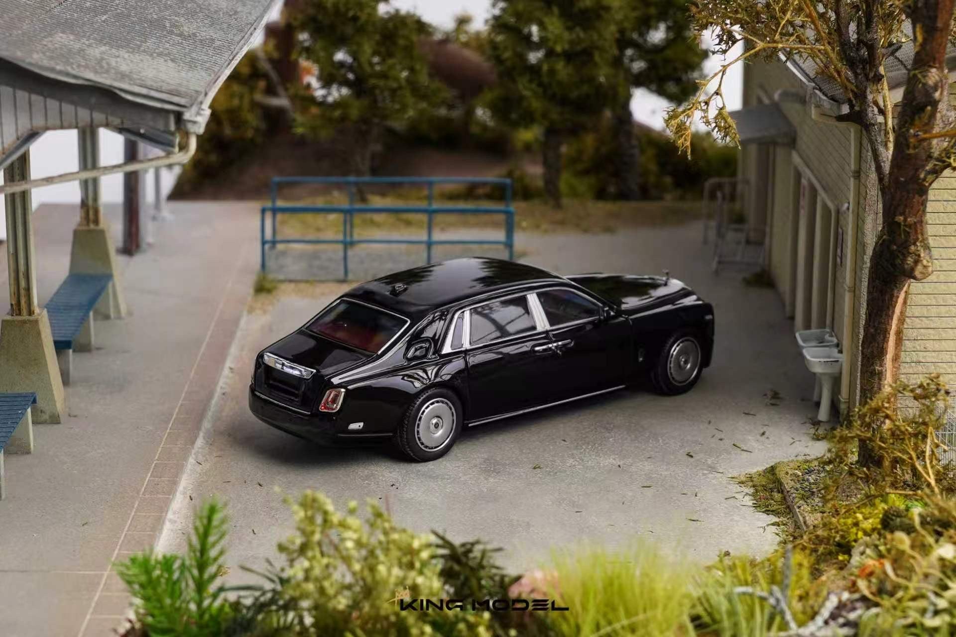 A black diecast model car resembling a luxury sedan, displayed on a miniature outdoor street scene with tiny buildings, trees, and a bench.