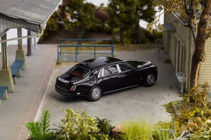A black diecast model car resembling a luxury sedan, displayed on a miniature outdoor street scene with tiny buildings, trees, and a bench.