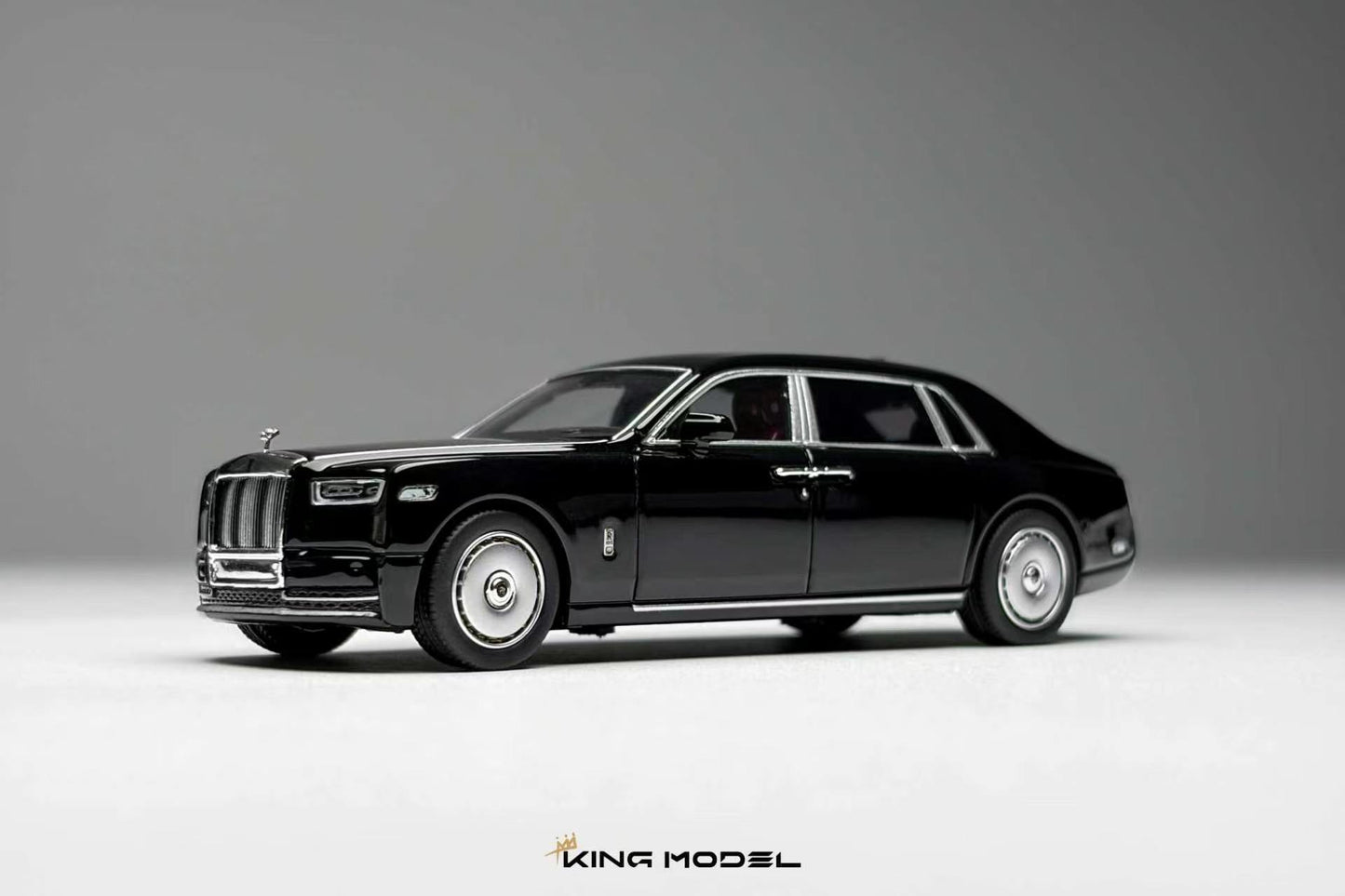 A black diecast model car of a luxury sedan, styled like a Rolls-Royce, shown from a three-quarters front angle on a neutral gray studio background. The model features chrome wheels, silver trim, and a detailed grille.