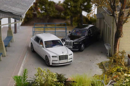 A miniature model diorama of a white Rolls-Royce Phantom and a black Rolls-Royce Cullion parked in a detailed model village street scene. The image is taken from an elevated angle, showing the scale models in a diorama setting with miniature houses, trees, and pathways.
