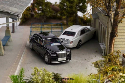A miniature model diorama of two luxury cars, a black Rolls-Royce Phantom and a white Rolls-Royce Spectre, parked in a detailed model village street scene.