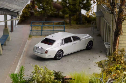 A white model car, resembling a Rolls-Royce Phantom, displayed in a detailed miniature diorama setting with a house, trees, and walkway.