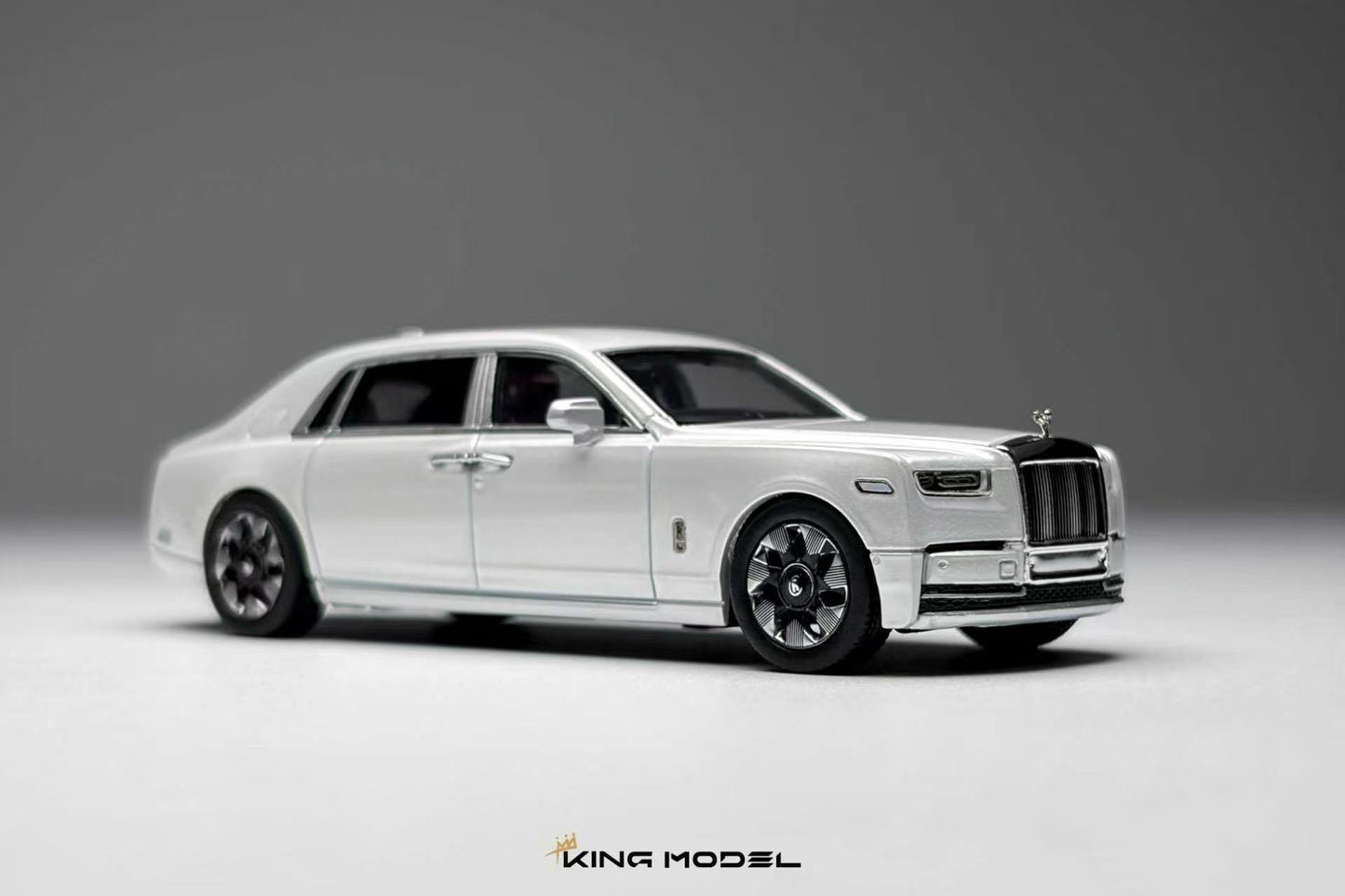 A white diecast model of a Rolls-Royce Ghost limousine, shown from a three-quarters front angle on a neutral gray studio background. The model features chrome details, black wheels, and a silver Spirit of London grille.