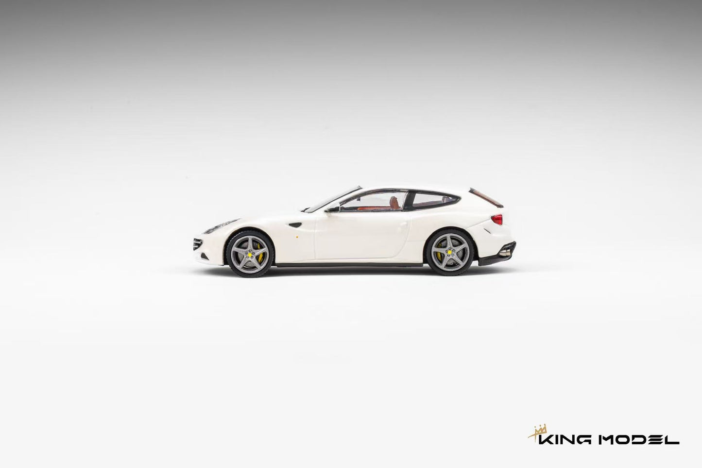 A white diecast model car, resembling a Ferrari FF, shown in a side profile on a light gray gradient background. The model features silver multi-spoke wheels and a small red accent on the rear side.