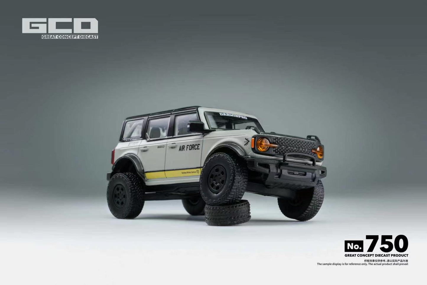 A scale model of a Ford Bronco in a light gray and white military livery with 'AIR FORCE' lettering and a yellow stripe, displayed on a gray studio background with a tire prop under its front bumper.