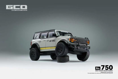 A scale model of a Ford Bronco in a light gray and white military livery with 'AIR FORCE' lettering and a yellow stripe, displayed on a gray studio background with a tire prop under its front bumper.