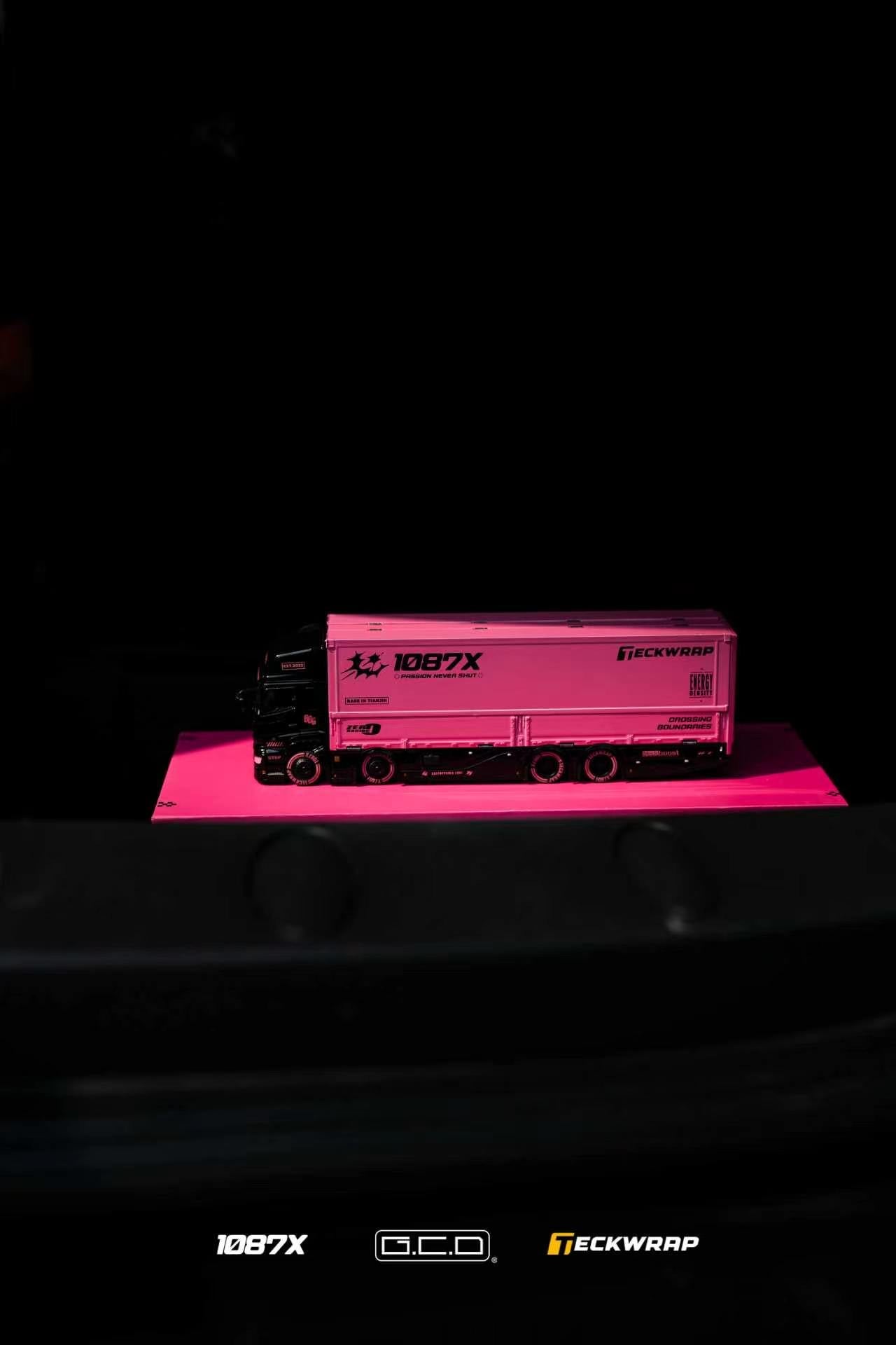 A pink semi-truck trailer model with black cab and printed branding, displayed on a bright pink platform against a dark background.