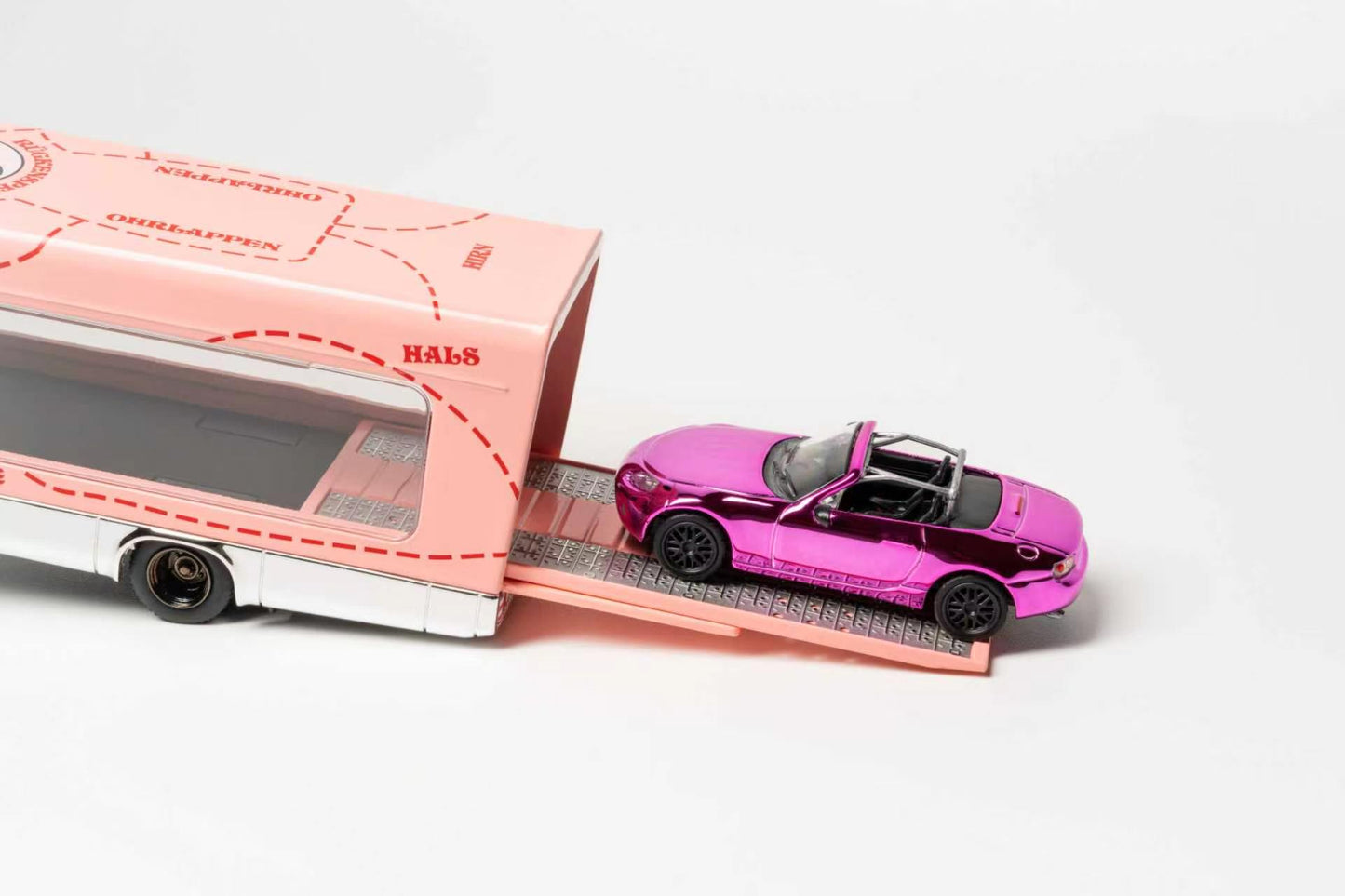 A pink toy convertible car with a black roof is shown on a ramp beside a pink and white toy trailer or bus, photographed on a clean white studio background.