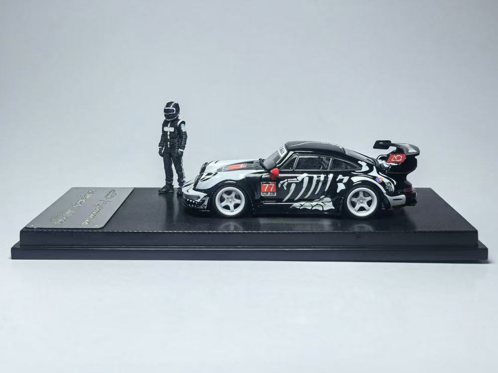 Miniature model of a black race car with white patterns and a red wing, placed on a display stand beside a small figure in a racing suit and helmet.