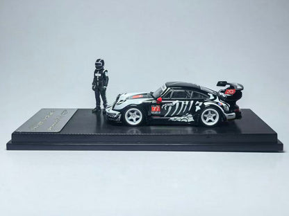 Miniature model of a black race car with white patterns and a red wing, placed on a display stand beside a small figure in a racing suit and helmet.