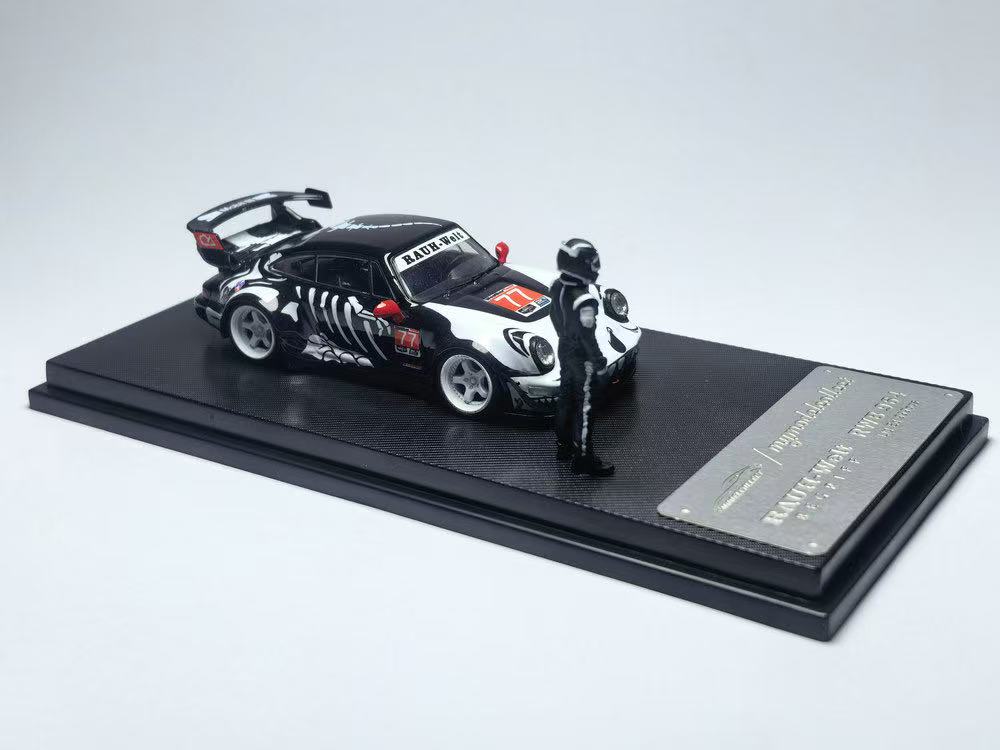 A miniature black race car with white decals and a red number 77 is displayed on a stand alongside a small helmeted driver figurine.