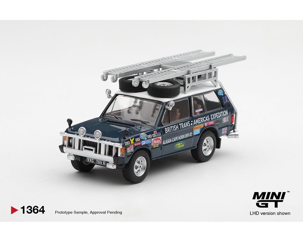 A miniature model of a vintage British Trans-Americas Expedition LHD car, shown on a clean light gray background. The model features a dark blue body, a silver roof rack with two spare tires, and various colorful racing decals.