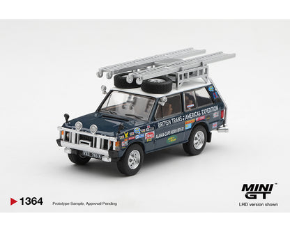 A miniature model of a vintage British Trans-Americas Expedition LHD car, shown on a clean light gray background. The model features a dark blue body, a silver roof rack with two spare tires, and various colorful racing decals.