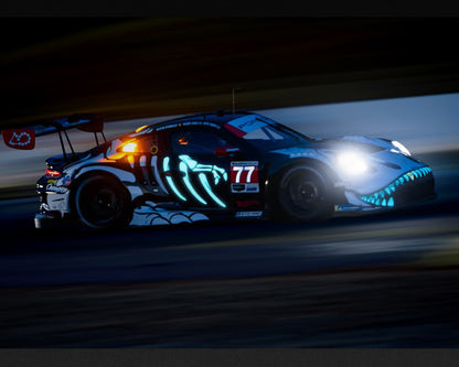 AO Racing Porsche 911 GT3 R (992) #77 Skeleton Rexy racing at night during the 2026 IMSA Petit Le Mans, featuring glowing blue dinosaur skeleton graphics, illuminated teeth, headlights and black-and-white racing livery. Reference image for Mini GT 1:64 MGT01436-BL.