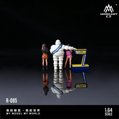 Model figure of a Michelin man with two people and a game console, on a reflective surface with 'MoreArt' branding.