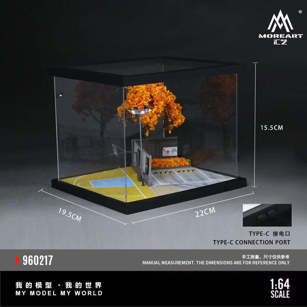 A miniature basketball court model displayed inside a clear acrylic display box with black trim. The model features a gray court surface with yellow and blue painted areas, a small bench, and a bright orange tree with matching orange leaves.