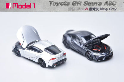 Two toy models of Toyota GR Supra cars in silver and navy gray on a light gray background.