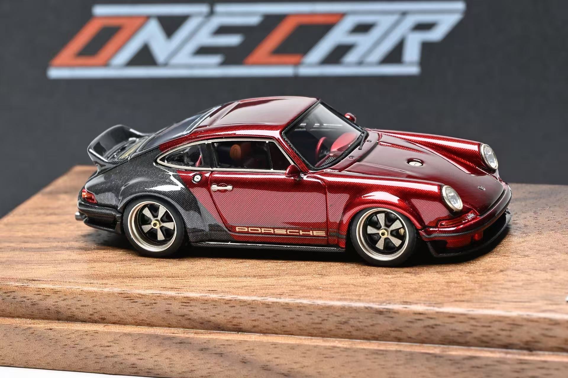 A scale model of a red and black Porsche 911 car displayed on a wooden base with a dark background and a 'ONE CAR' logo.
