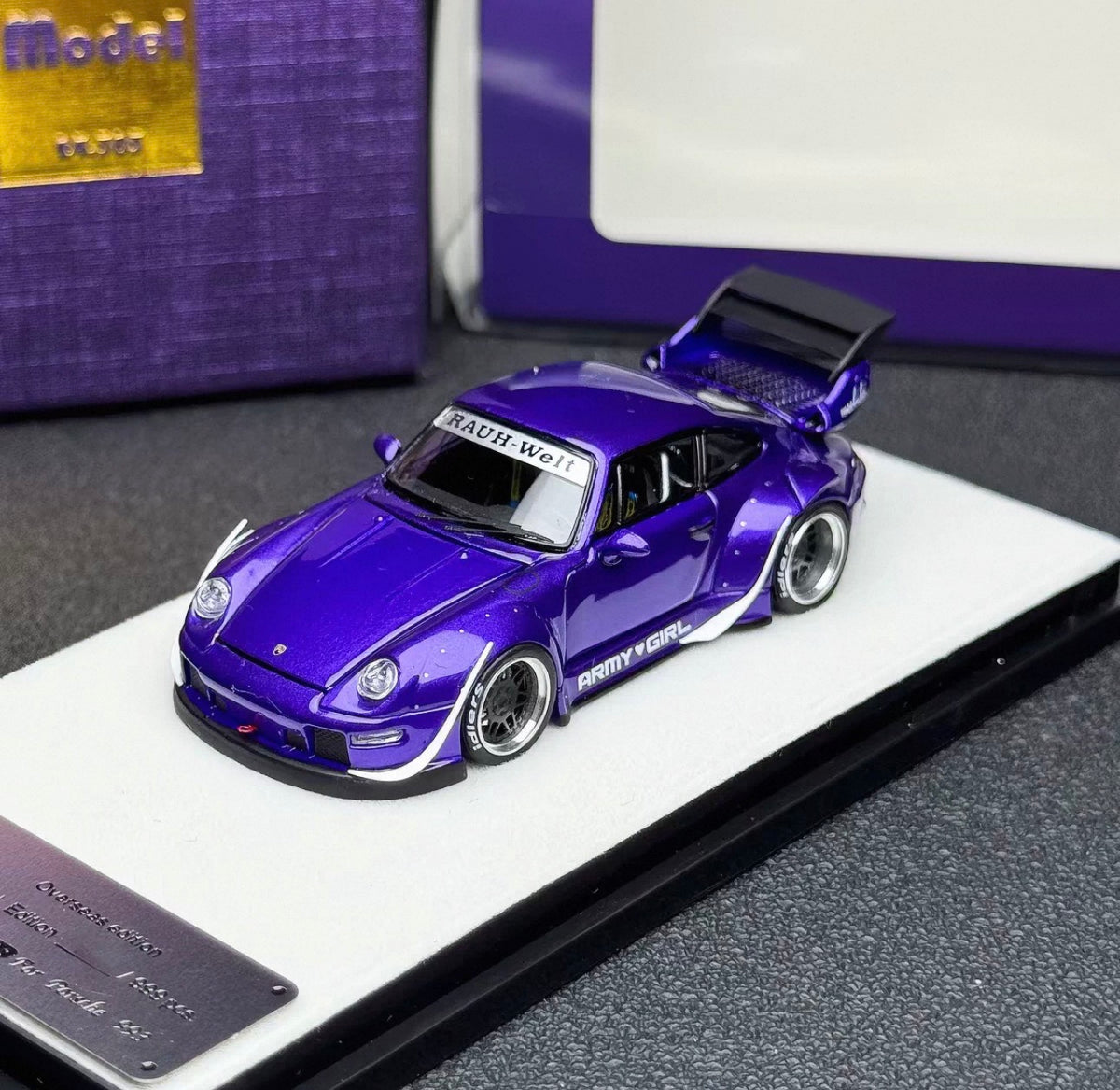 A purple widebody model car displayed on a white stand, with a purple box in the background and a small badge plaque in the foreground.