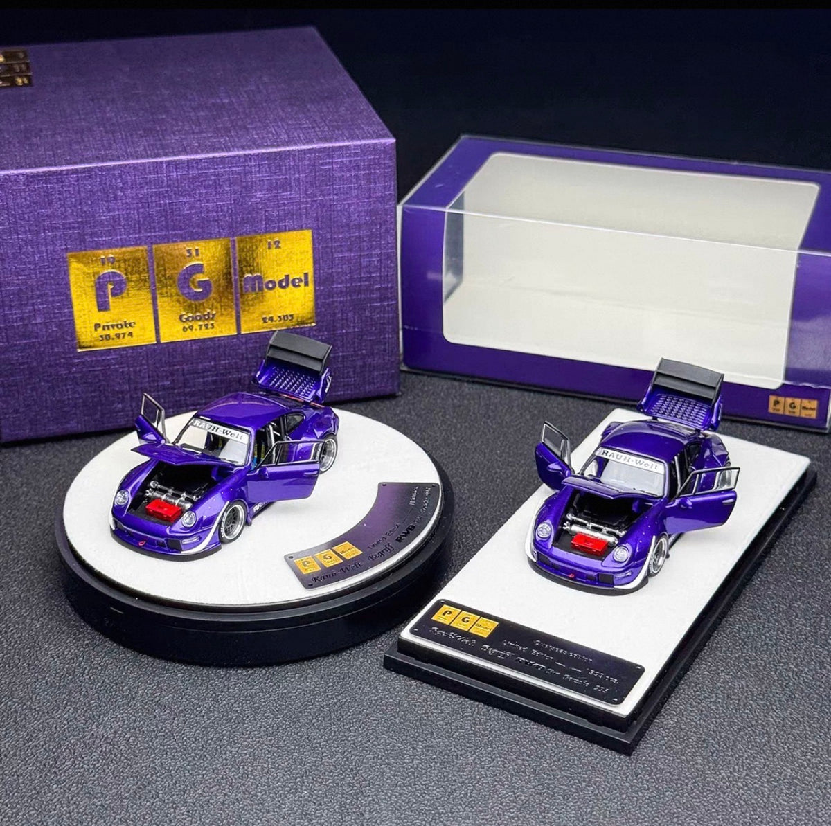 Two miniature purple model cars displayed on round and rectangular stands, with a purple branded box and a clear display box in the background.