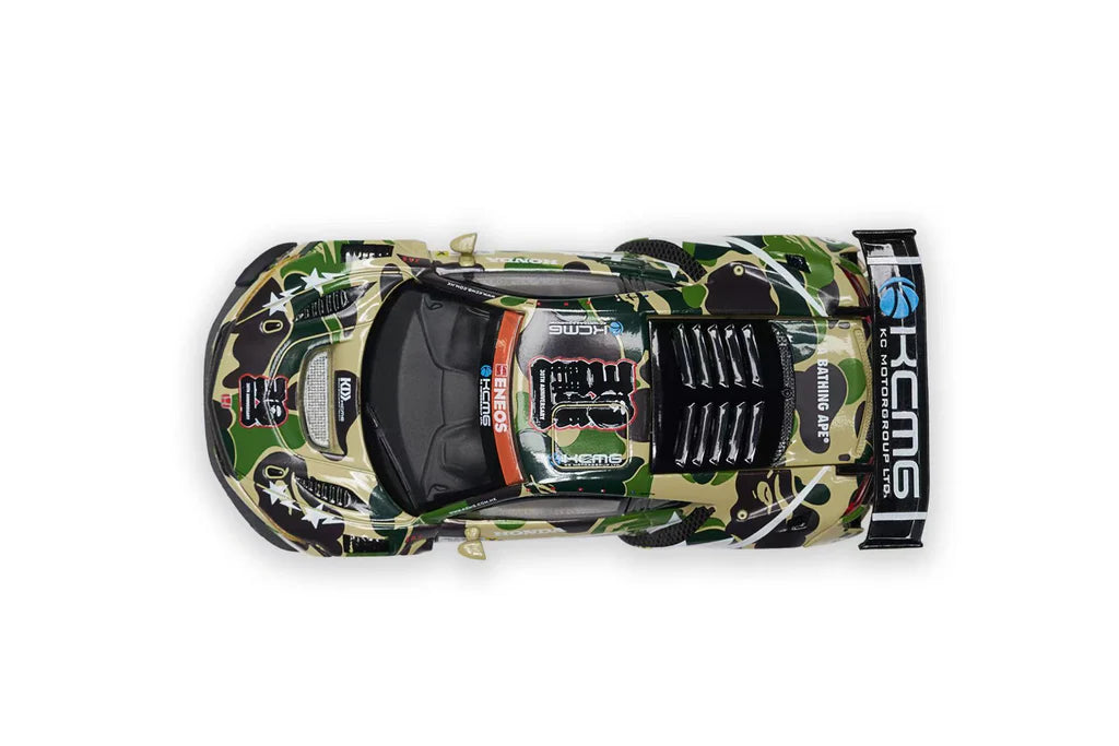 Top view of a race car with a green camo and black pattern, branded with the logos "KC MG" and "BATHING APE," evoking speed and intensity.