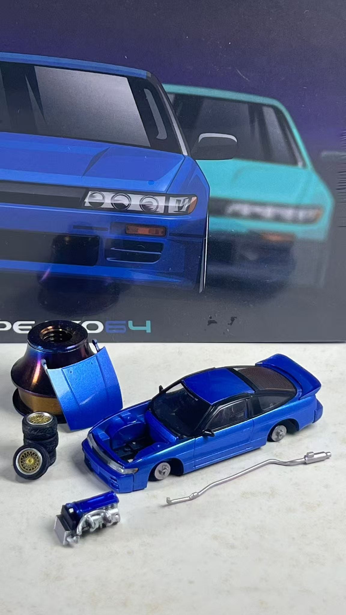 A blue diecast model car shown disassembled with various parts laid out on a light surface, with its original packaging box in the background.