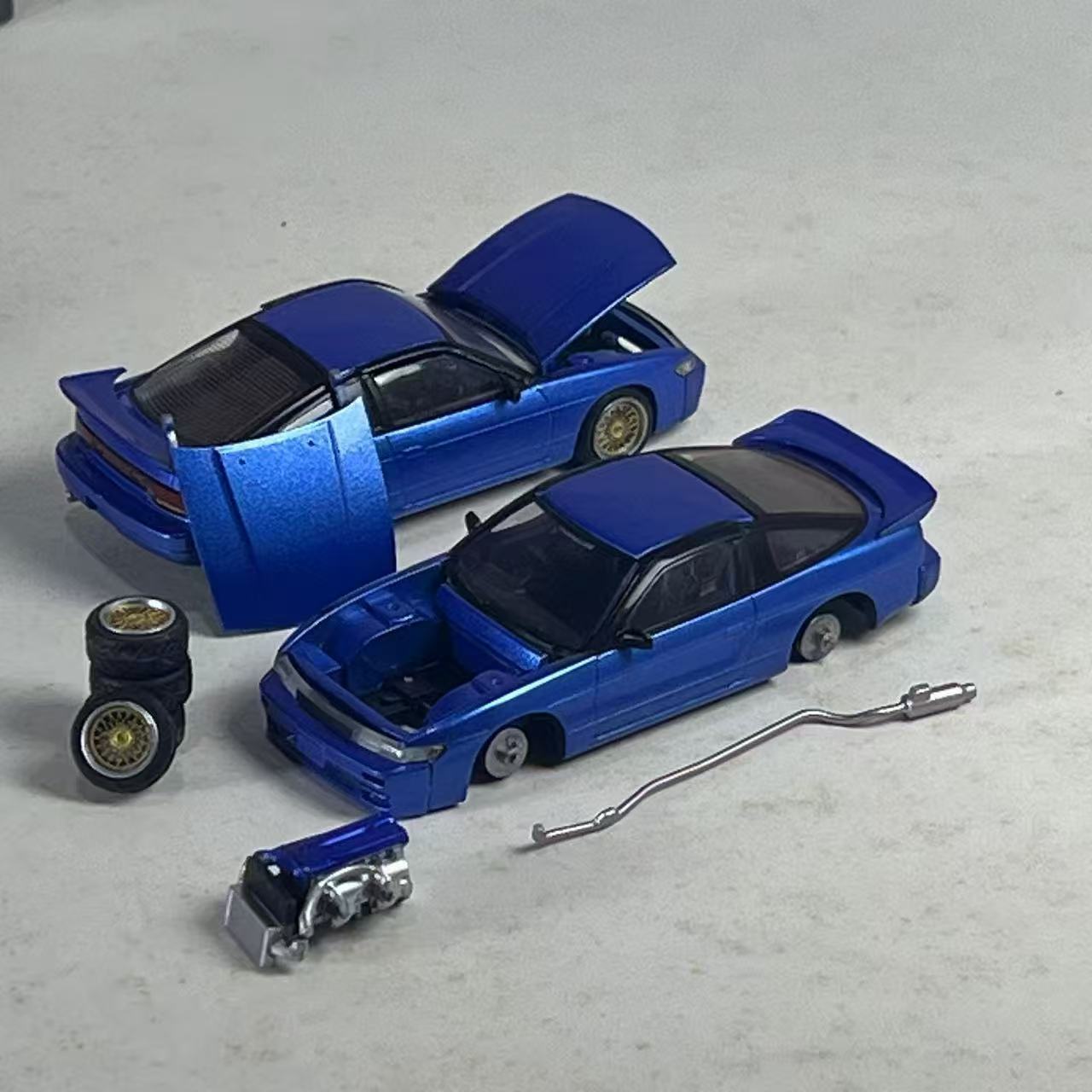 Two metallic blue die-cast toy cars with open hoods, shown on a light gray surface with small accessory parts including wheels, a metal bar, and a small blue mechanical piece.