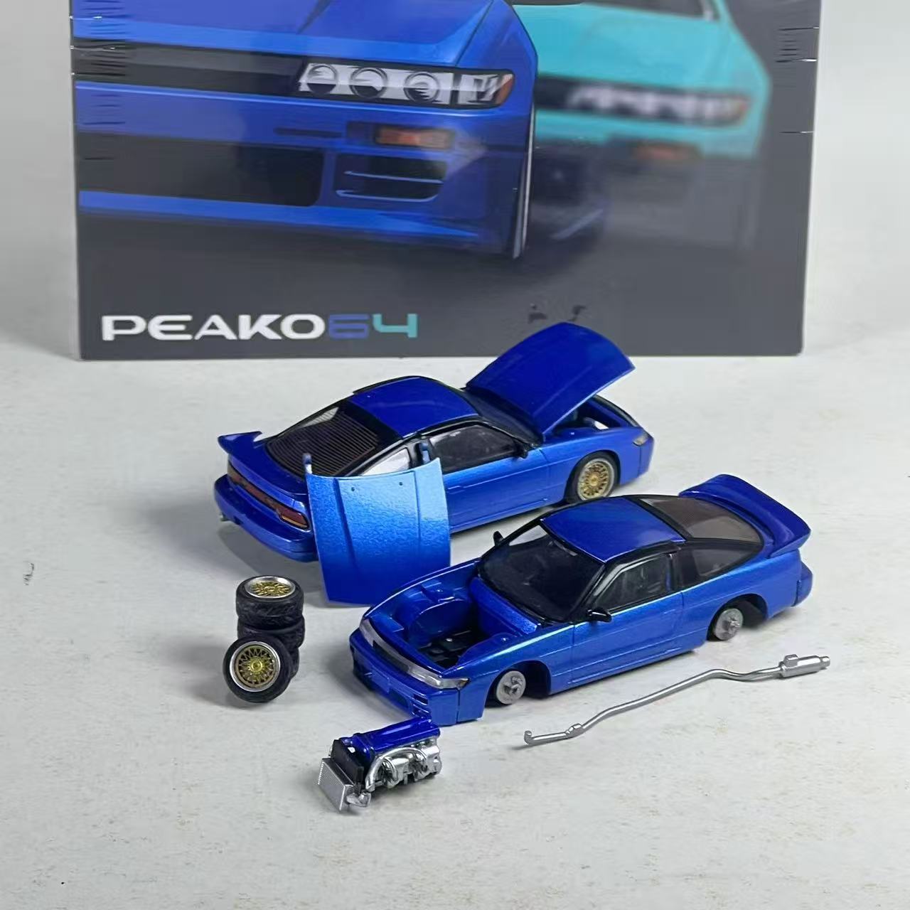 Two metallic blue die-cast model cars with open hoods and doors, displayed with small accessories including wheels, a long metal rod, and a miniature engine on a light gray surface, with the branded product box in the background.