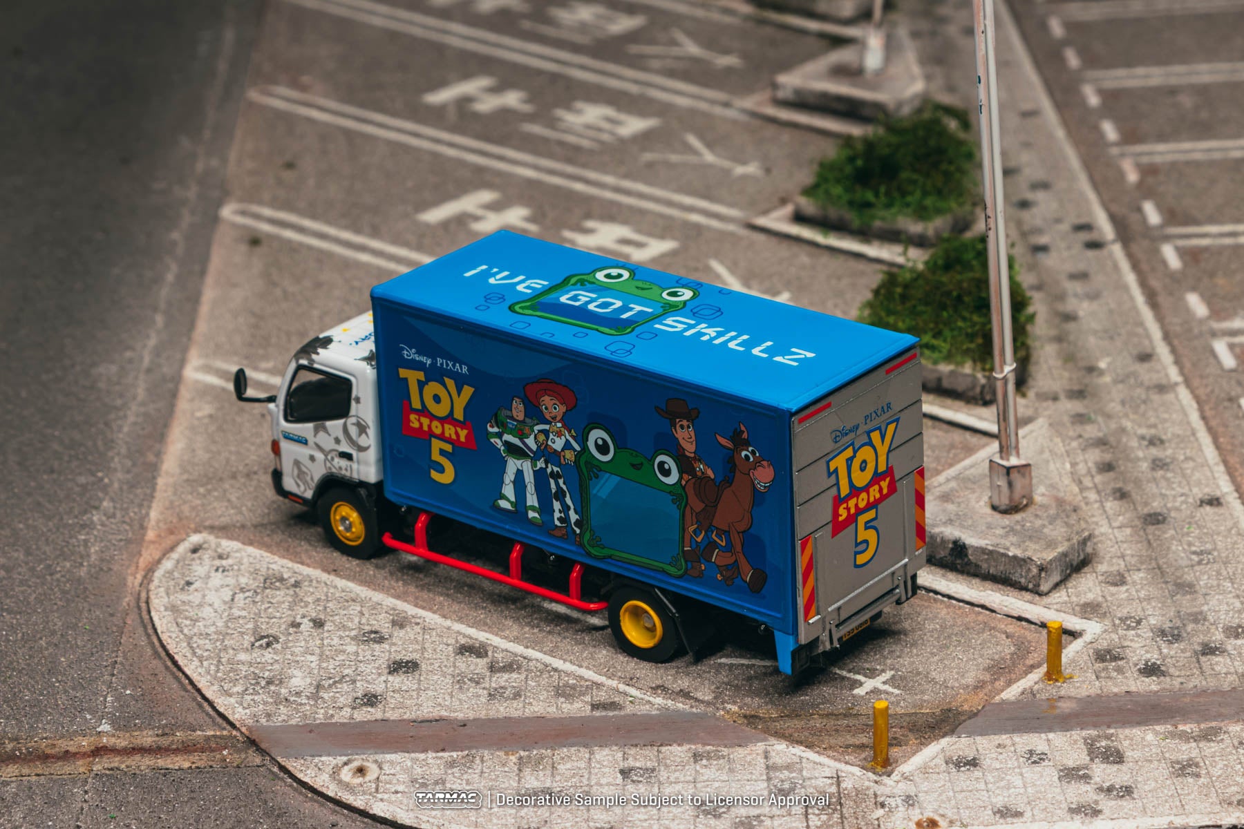 A diecast model toy truck featuring a blue Toy Story 5 container with characters Woody, Buzz Lightyear, Jessie, and Bullseye, parked on a miniature street intersection.