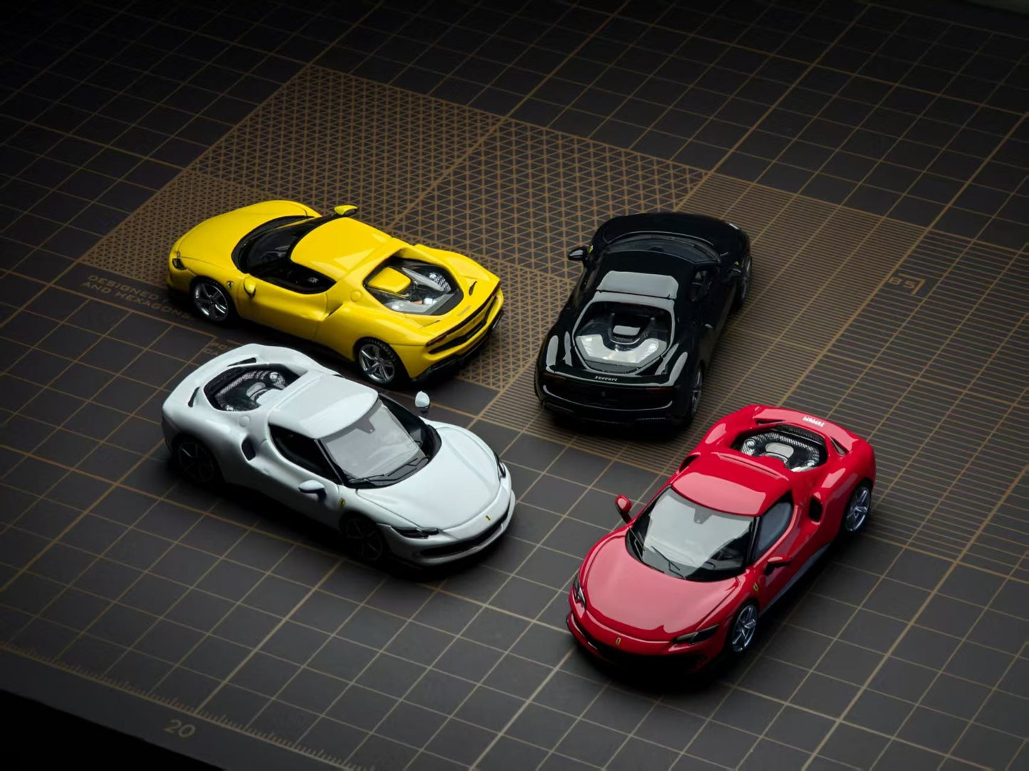 Four miniature toy sports cars in yellow, black, white, and red, arranged on a dark grid-patterned surface.