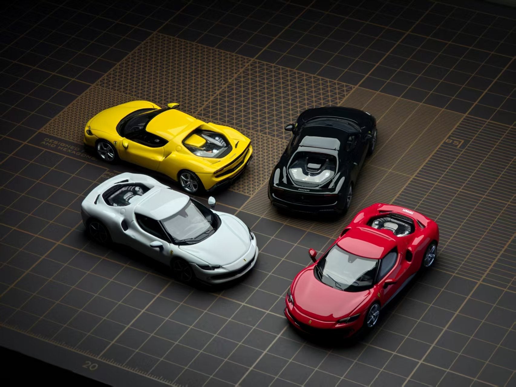 Four miniature toy sports cars in yellow, black, white, and red, arranged on a dark grid-patterned surface.
