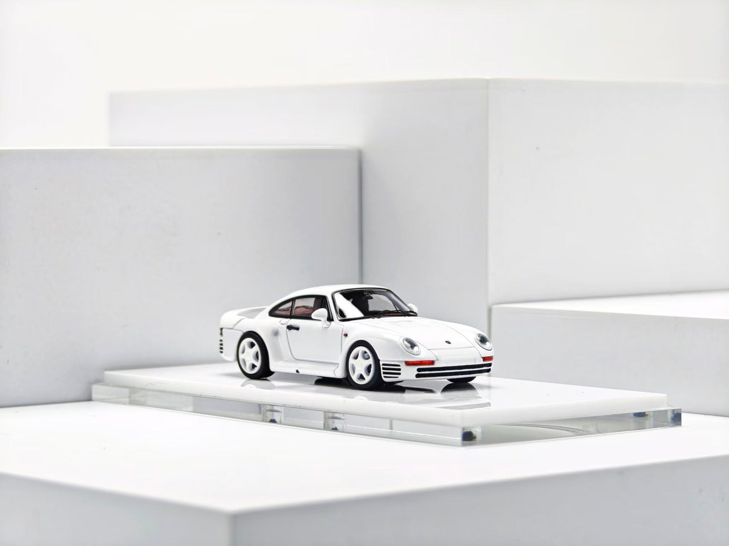 A miniature white Porsche 911 model car displayed on a clear acrylic platform against a minimalist white geometric background.