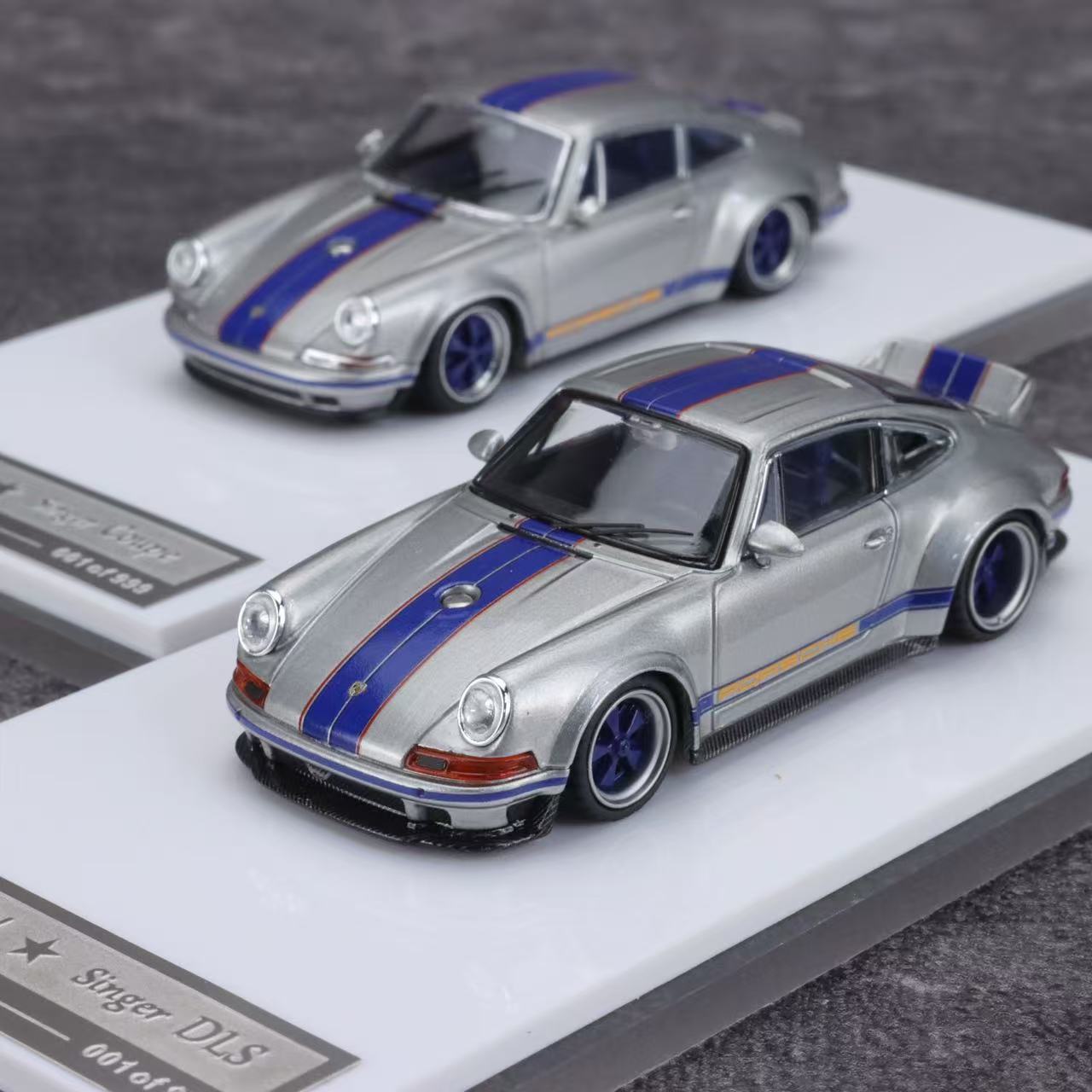 Two model cars, a silver and blue Porsche with stripes, on display stands.