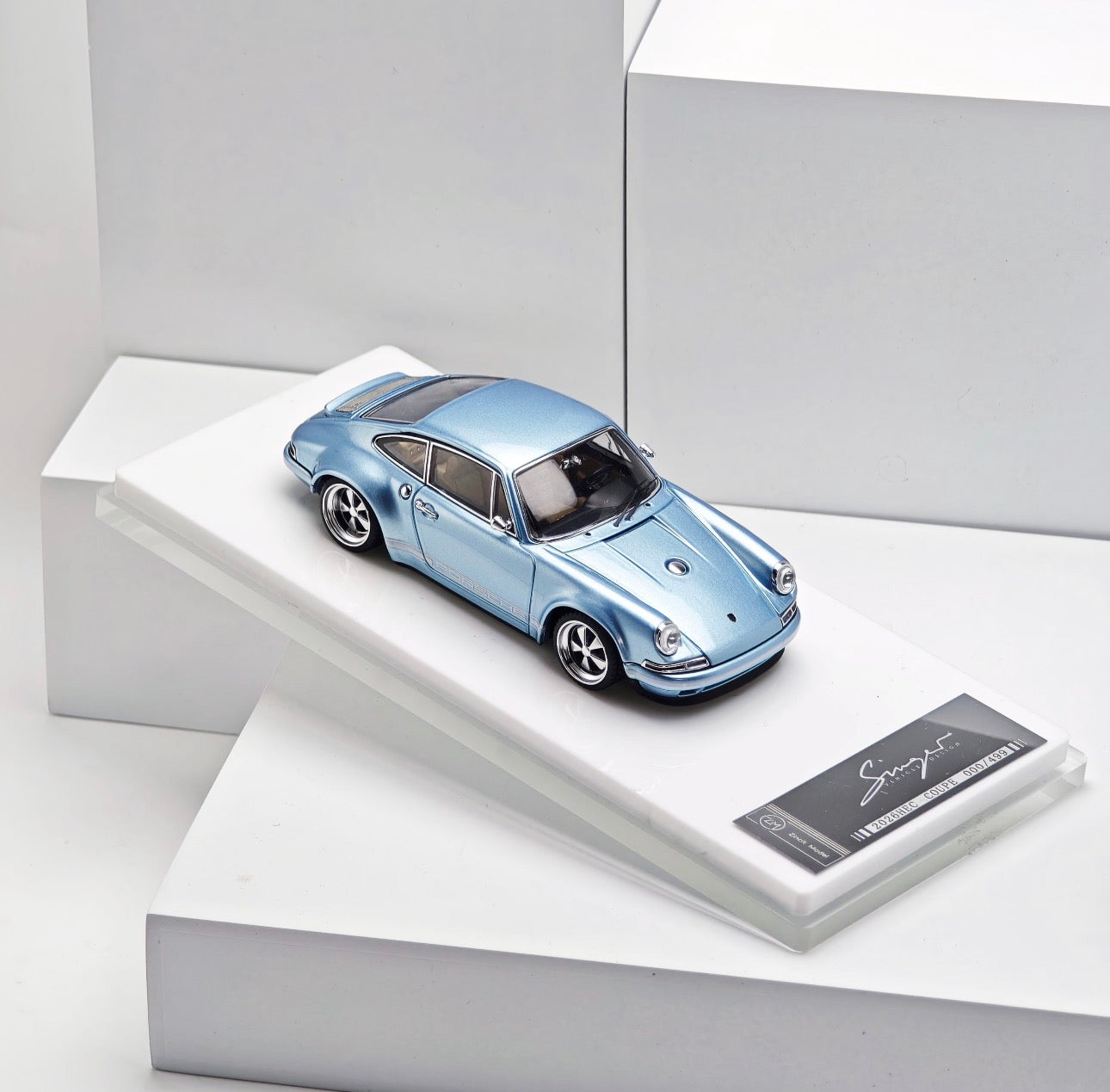 A metallic light blue model car displayed on a white angled pedestal with a small black plaque, photographed in a clean white studio setting.