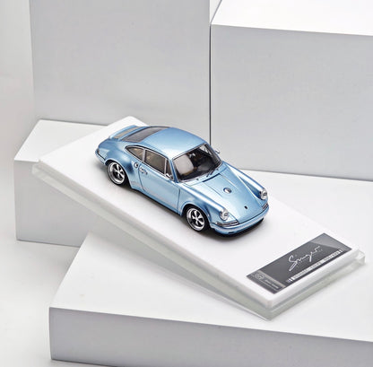 A metallic light blue model car displayed on a white angled pedestal with a small black plaque, photographed in a clean white studio setting.