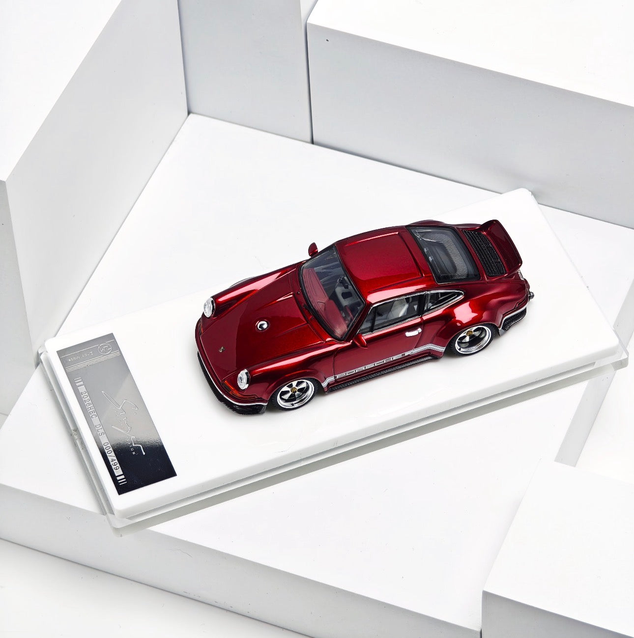 A metallic red Porsche 911 model car displayed on a white rectangular base with a small product card, photographed in a clean white geometric setting.